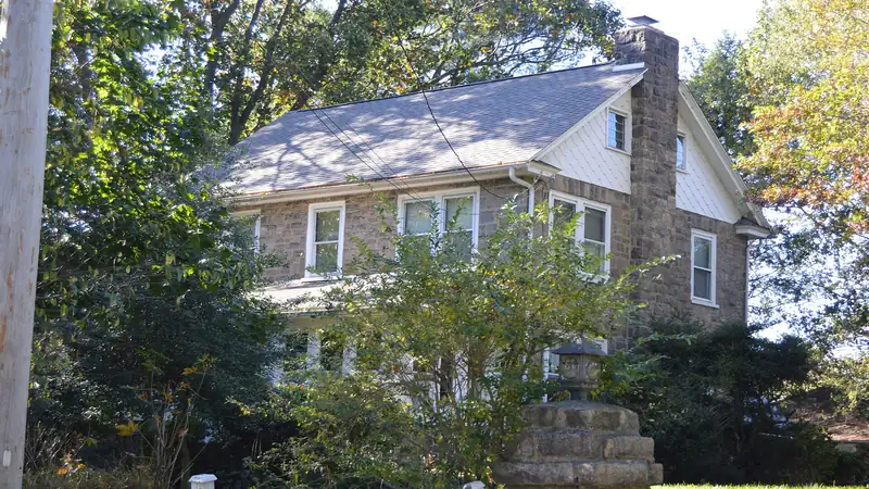 Two-story stone house with a gabled shingle roof, surrounded by trees